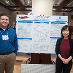 Two student presenters standing on either side of their shared psoter, smiling and posing for a photo